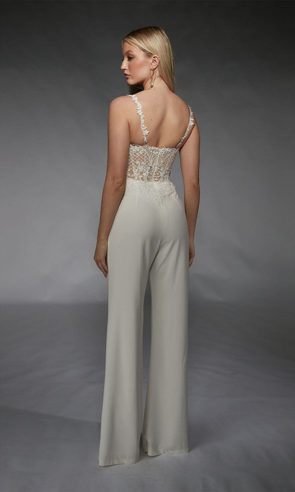 A woman faces away in the Alyce 70074 Long Formal Jumpsuit, featuring a white design with lace and floral embroidery on the back and wide-leg pants, set against a plain gray background.